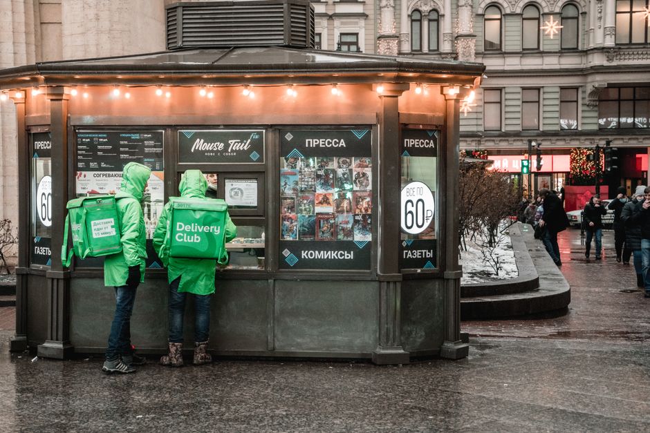 Two delivery couriers in green jackets stand by a food stall at night.